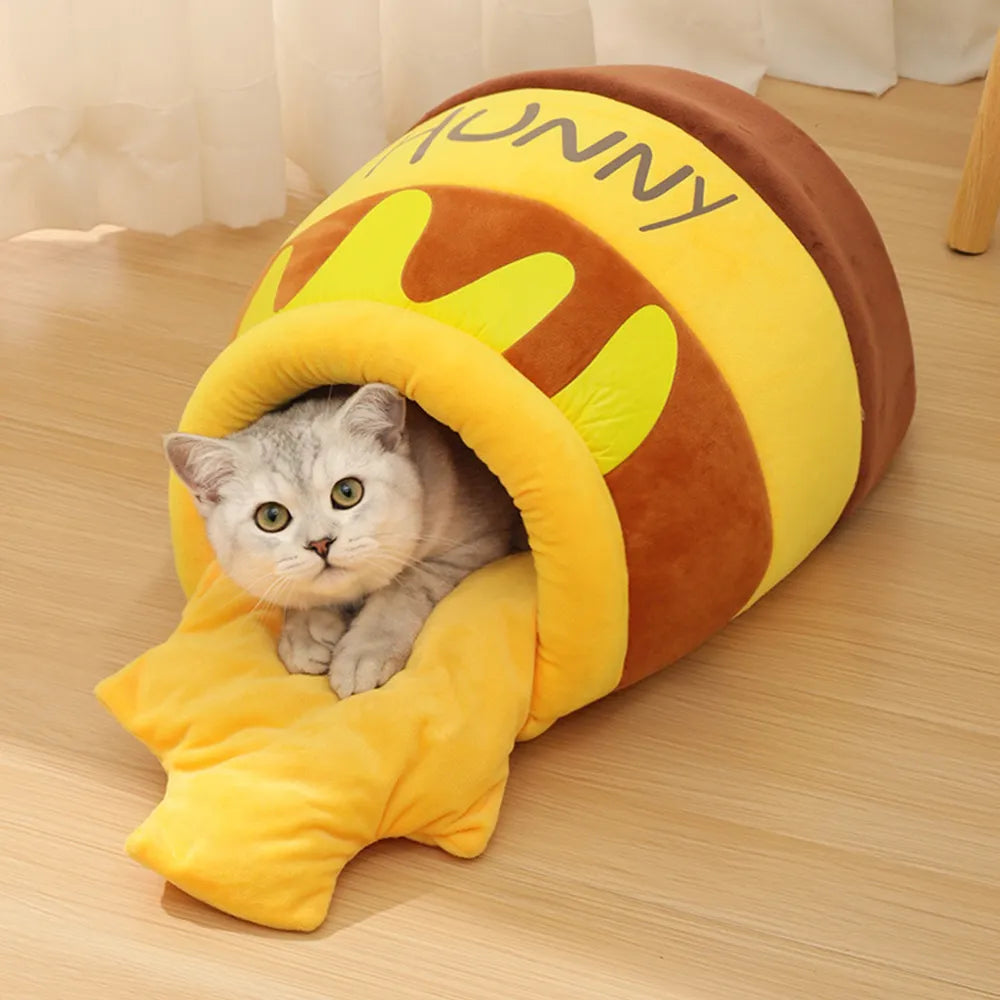 Gray cat resting inside yellow honey jar–themed plush cat cave with blanket.